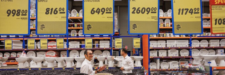 A store display showing various sanitary items and price tags, with two employees interacting in the foreground.
