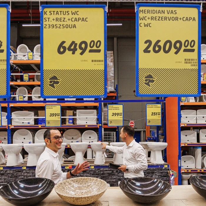 A store display showing various sanitary items and price tags, with two employees interacting in the foreground.