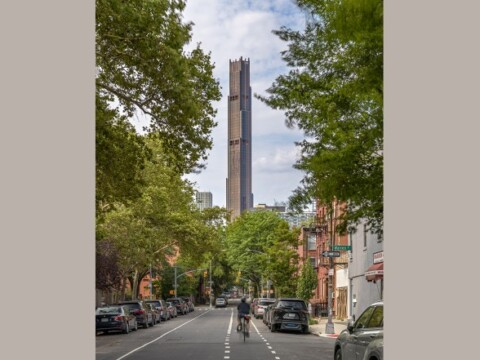 A street lined with trees and parked cars, featuring a tall tower in the background under a cloudy sky.