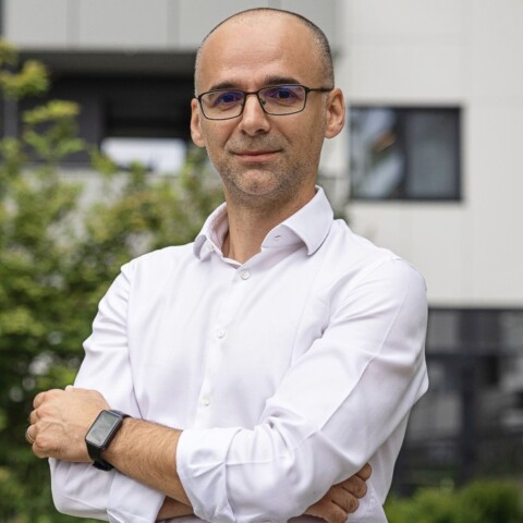 A man in a white shirt stands outdoors with arms crossed, in front of a building and greenery.