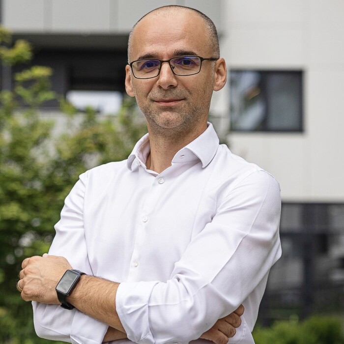 Mann mit Brille und Hemd, steht mit verschränkten Armen vor einem modernen Gebäude und Bäumen.