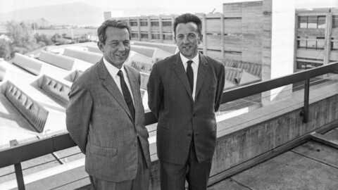 Zwei Männer in Anzug stehen lächelnd auf einem Balkon mit moderner Architektur im Hintergrund.