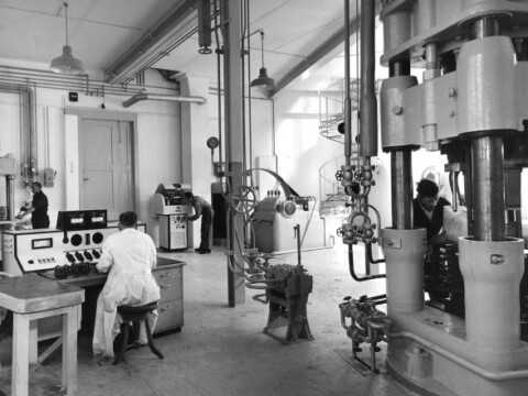 Ein industrieller Arbeitsplatz mit mehreren Maschinen und Geräten, Techniker in Laborkitteln arbeiten an Steuerpulten und Apparaturen.