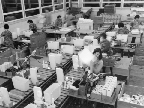 Ein Grossraumbereich mit mehreren Arbeiterinnen und Arbeitern an Montagelinien, die verschiedene Plastikprodukte assemblieren.