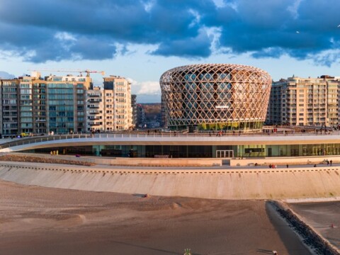 Modern architectural structure with a distinctive circular design set against a backdrop of residential buildings and a sandy shoreline.