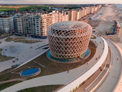 A modern, round building with a geometric facade surrounded by grassy areas and a walkway, overlooking a beach and coastal cityscape.