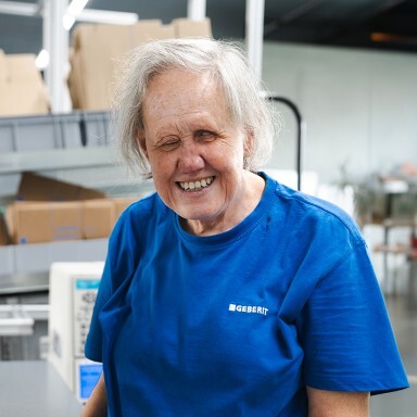 Eine lächelnde ältere Frau mit grauen Haaren trägt ein blaues T-Shirt und steht an einem Tisch in einem Werkstatt- oder Lagerumfeld.