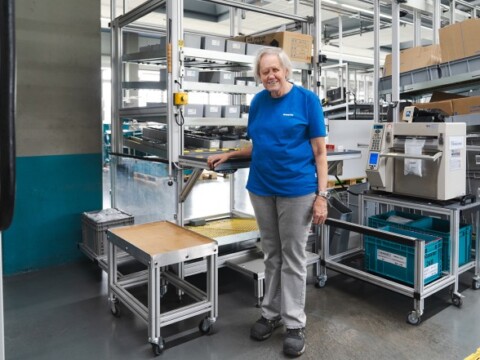 Eine lächelnde Frau in einem blauen T-Shirt steht neben einem mobilen Regal mit einer Arbeitsfläche und anderen Lagerregalen in einer modernen Fabrik.