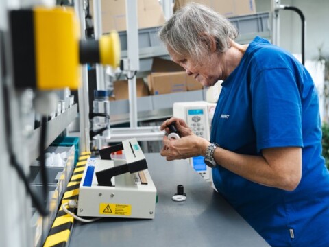 Eine ältere Frau in einem blauen T-Shirt arbeitet an einem Tisch, während sie ein kleines Objekt in der Hand hält und ein Gerät bedient.