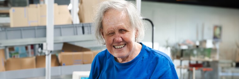 Eine lächelnde ältere Person mit grauen Haaren trägt ein blaues T-Shirt und steht in einer hellen, modernen Umgebung mit Kartons im Hintergrund.