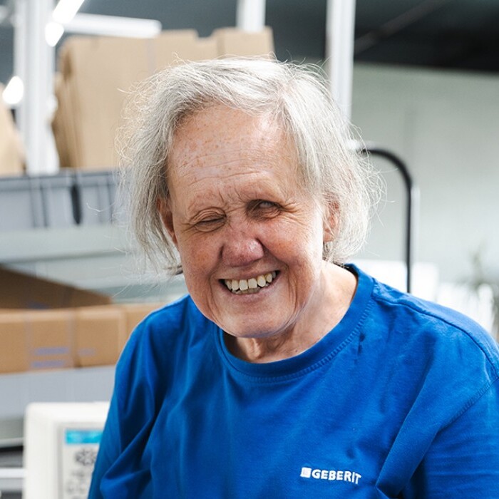 Eine lächelnde ältere Person mit grauen Haaren trägt ein blaues T-Shirt und steht in einer hellen, modernen Umgebung mit Kartons im Hintergrund.