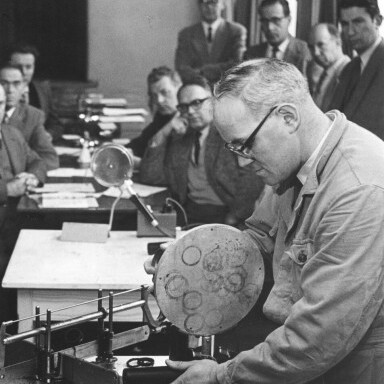 Ein Mann demonstriert eine technische Maschine vor einer Gruppe von Zuschauern in einem Schulungsraum.