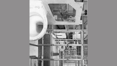 Eine technische Ansicht einer Toilette mit sichtbaren Rohrleitungen und einem gitterartigen Gestell um sie herum.