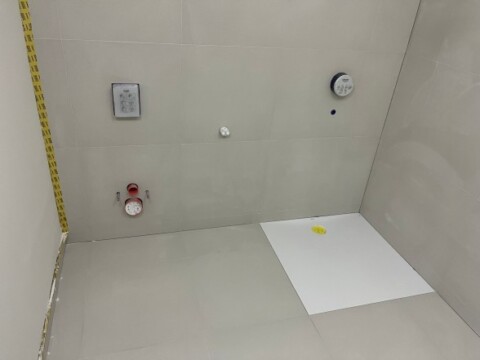 A tiled wall with plumbing fixtures, including a shower head and temperature control, and a section of the floor with markings.