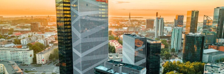 City skyline at sunset featuring modern high-rise buildings, including a prominent glass tower with a geometric pattern.