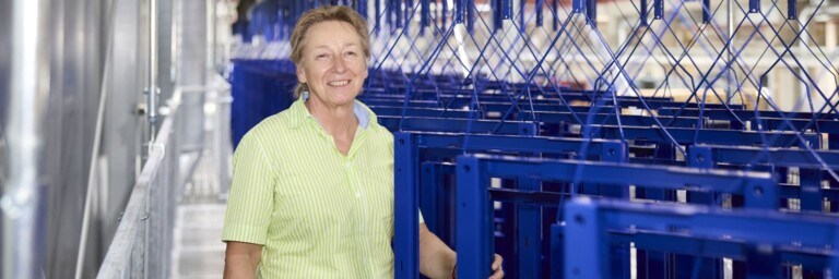 A smiling woman in a green shirt stands beside blue metal frames in a warehouse setting.