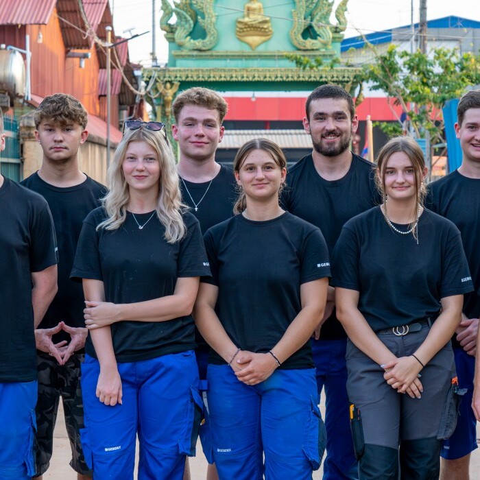 A diverse group of twelve young individuals stands in front of a colorful building, all wearing black t-shirts and blue pants.