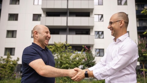 Zwei Männer schütteln sich die Hände vor einem modernen Wohngebäude, beide lächeln freundlich.