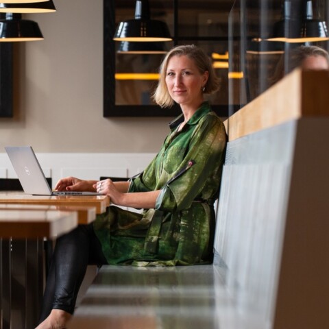 Eine Frau sitzt in einem modernen Brasserie auf einer Bank, mit einem Laptop vor sich und lächelt in die Kamera, während im Hintergrund Tische und Lampen sichtbar sind.