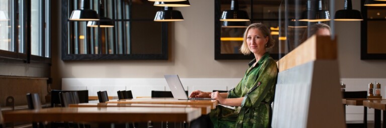 Eine Frau sitzt in einem modernen Brasserie auf einer Bank, mit einem Laptop vor sich und lächelt in die Kamera, während im Hintergrund Tische und Lampen sichtbar sind.