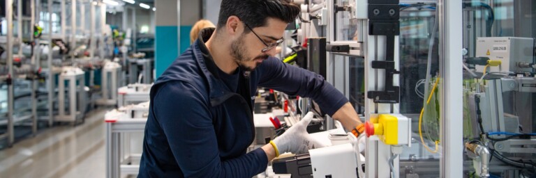 Ein Fabrikarbeiter in einem modernen Produktionsraum montiert ein Gerät mit einer präzisen Handbewegung.