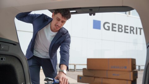A man in a blazer leans into the trunk of a car, looking at packages while a building with a Geberit sign is in the background.