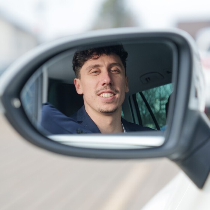 A smiling man is looking at the camera from a car's side mirror, with a blurred parking lot visible behind him.