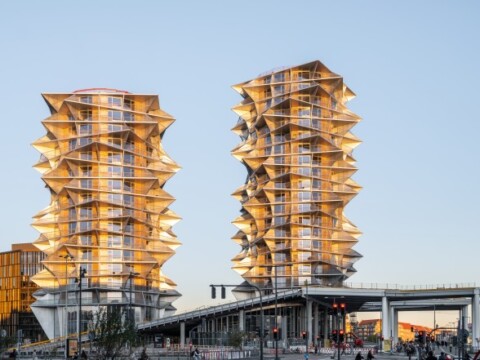 Zwei moderne Wolkenkratzer mit markanter, gewellter Fassade, in goldener Beleuchtung, umgeben von urbaner Landschaft.