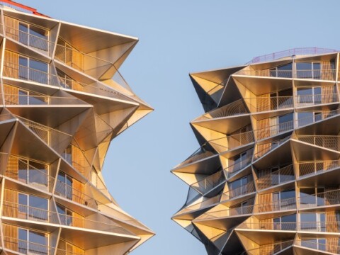 Zwei futuristisch gestaltete Wolkenkratzer aus Glas und Metall mit einer kantigen, schrägen Fassade, die im Sonnenlicht glänzen.