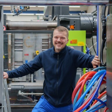 Mann in blauer Arbeitskleidung steht lächelnd zwischen Maschinen und bunten Schläuchen in einer industriellen Umgebung.