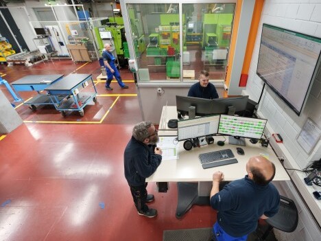 Inside a manufacturing facility, two workers discuss data at a desk with multiple monitors, while another person walks in the background.