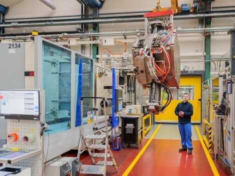 A man stands in a manufacturing facility beside large machinery, including a robotic arm and control panels.