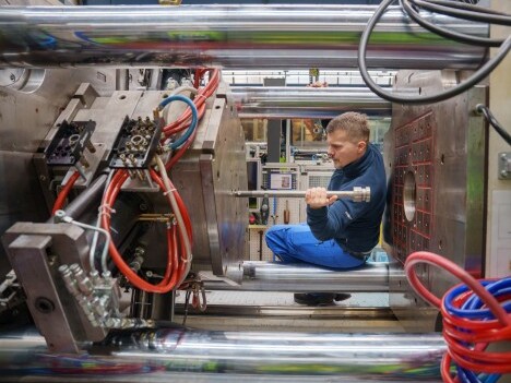 A man in blue overalls is seated inside a large industrial machine, working with metal components and colorful tubes.