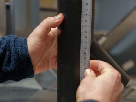 A person is measuring a black pipe with a ruler in a workshop setting.