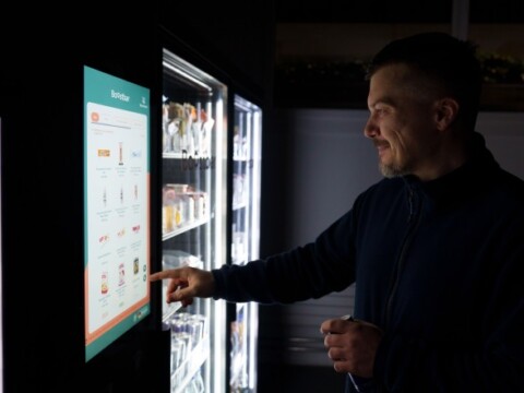 A man interacting with a touchscreen vending machine displaying various products in a dimly lit environment.