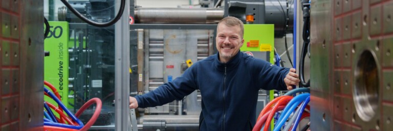 A smiling man in a blue jacket stands between large machines, surrounded by colourful hoses in an industrial setting.