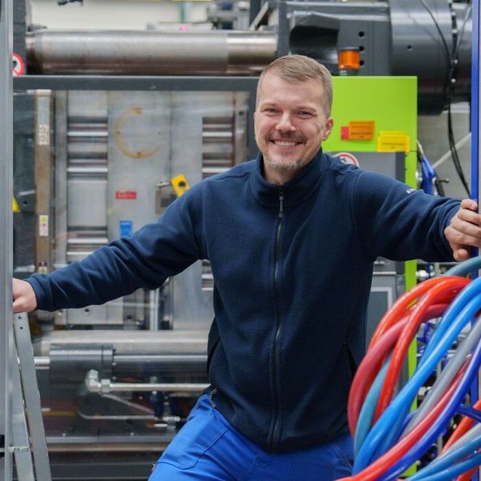 A smiling man in a blue jacket stands between large machines, surrounded by colourful hoses in an industrial setting.