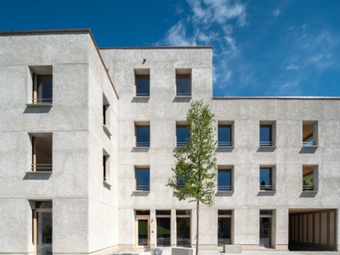 Modern, minimalistisch gestaltetes Gebäude mit Betonfassade und mehreren Fensterreihen, umgeben von klar blauem Himmel.