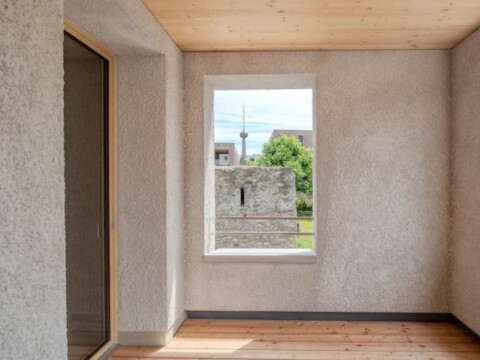 Heller Innenraum mit Holzfussboden und Fensterblick auf einen Turm und eine grüne Landschaft.