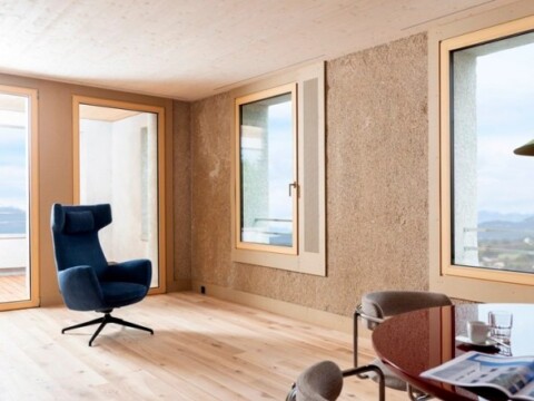 Ein moderner, heller Innenraum mit Holzfussboden, einem blauen Sessel und grossen Fenstern, die Sicht auf eine Landschaft bieten.