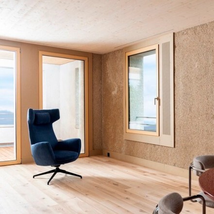A modern interior featuring a blue armchair, large windows with wooden frames, and wooden flooring.