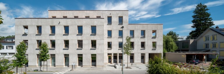 Modernes, mehrstöckiges Gebäude mit grauer Fassade und grossen Fenstern, umgeben von Bäumen und Rasenflächen.