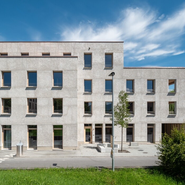 Modernes, mehrstöckiges Gebäude mit grauer Fassade und grossen Fenstern, umgeben von Bäumen und Rasenflächen.