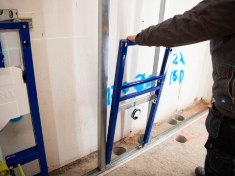 Eine Person montiert ein blaues Metallgerüst an einer Wand, während eine grüne Laserlinie sichtbar ist.