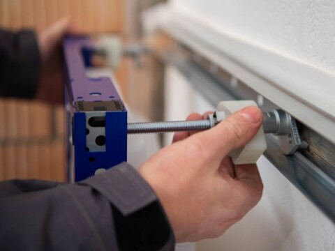 Eine Person installiert ein blaues Bauteil an einer Wand, während ein Gewindebolzen an einem Schienenmechanismus angepasst wird.