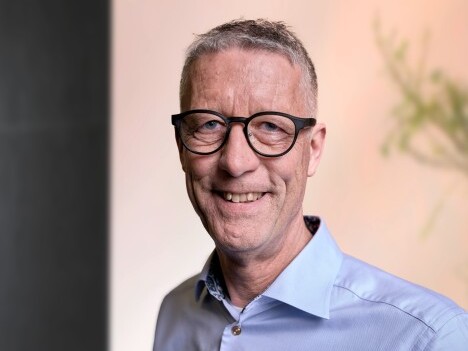A smiling middle-aged man with glasses, wearing a light blue shirt, standing in front of a softly blurred background.