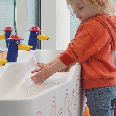 A young child in an orange hoodie washes hands at a playful sink with colorful spouts.