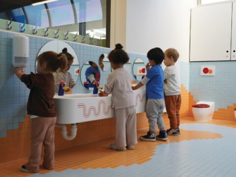 A group of four young children washing their hands at a low sink in a brightly colored bathroom.