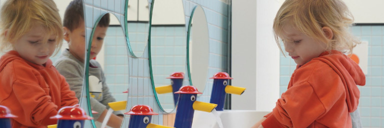 A child in an orange hoodie washes hands at a sink in a colorful bathroom, with playful faucet designs and mirrors.