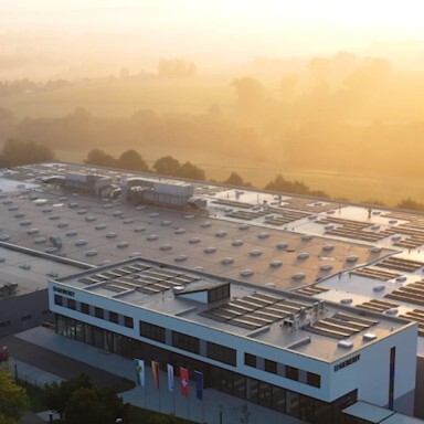 Aerial view of a modern building with a flat roof, surrounded by greenery and foggy landscapes during sunrise.